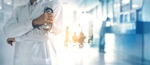 Keine Empathie für Ungeimpfte: Doktor im Kittel steht mit verschränkten Armen und im Hintergrund sind Menschen im Krankenhaus, offensichtlich krank