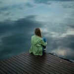 Alleinerziehend und einsam: eine junge Frau von hinten bei Regenwetter am See, dicke Wolken am verhangenen Himmel