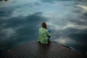 Alleinerziehend und einsam: eine junge Frau von hinten bei Regenwetter am See, dicke Wolken am verhangenen Himmel