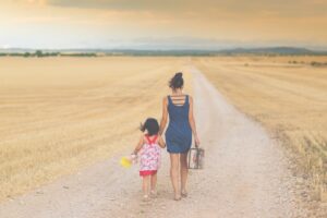 Mutter mit Koffer in der Hand und 4-jähriges Mädchen mit Teddybär in der Hand laufen auf einem Weg. Von hinten, in sonenkleider, am Horizont nur Landschaft, endloser Horizont