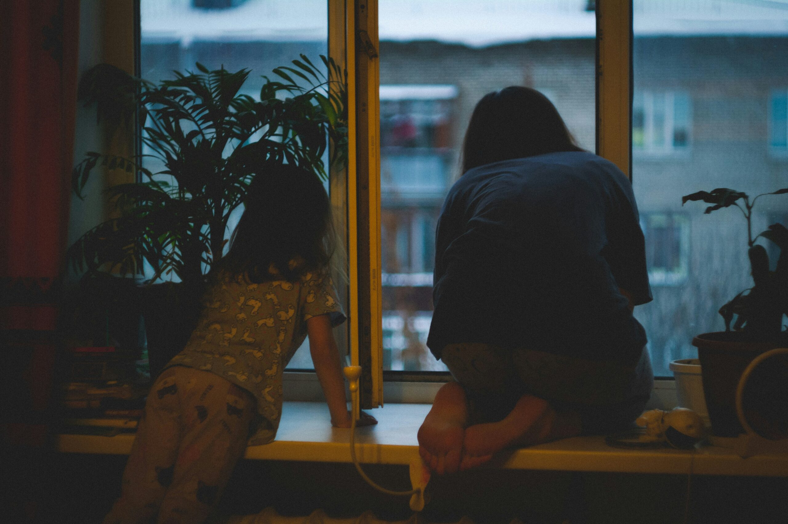 ein kind von hinten, es sitzt am fenster und schaut in die dämmerung einer stadt