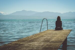 Introvertierte Frau sitzt allein auf einem Steg am See und blickt in Ruhe auf das Wasser und die Berge