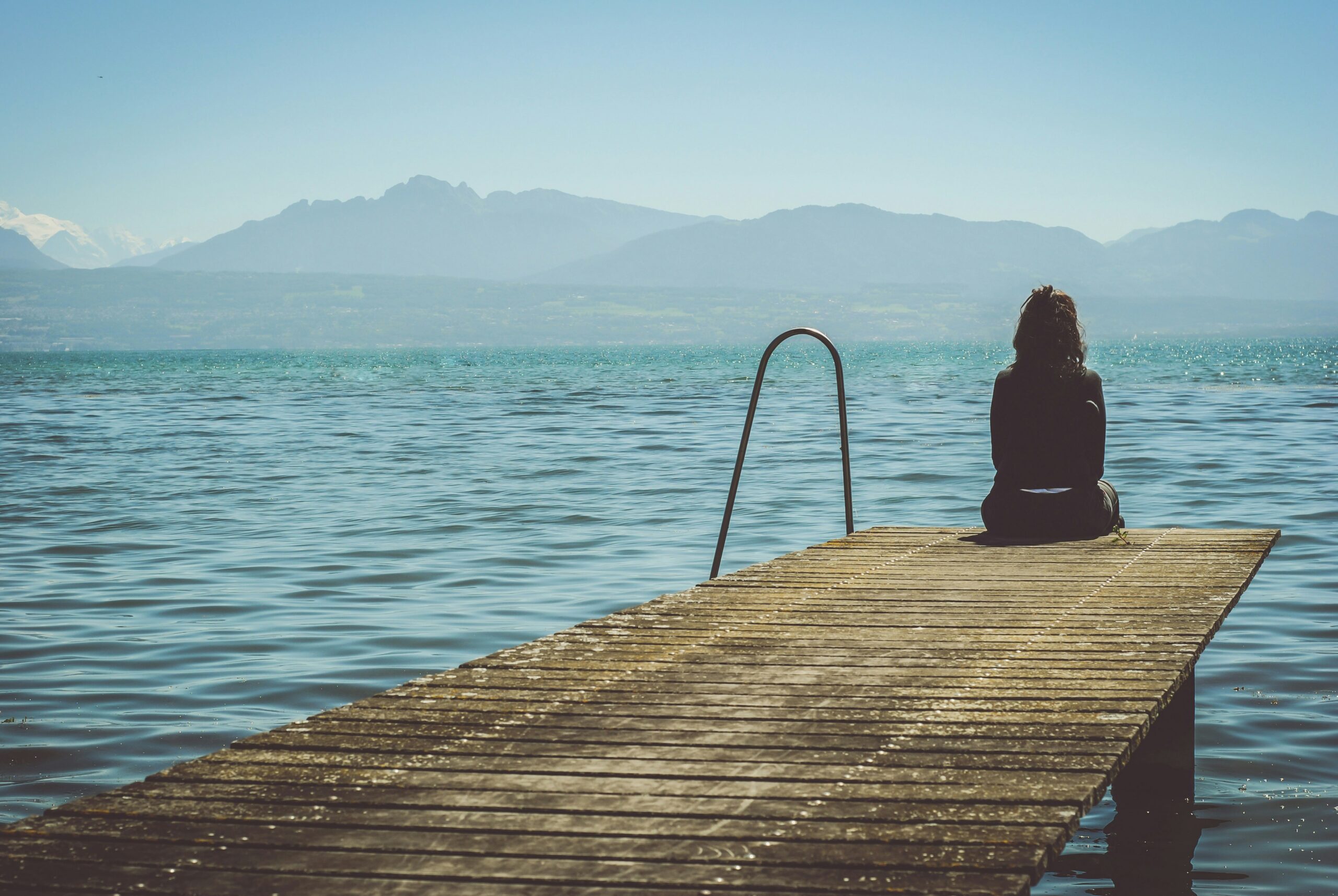 Introvertierte Frau sitzt allein auf einem Steg am See und blickt in Ruhe auf das Wasser und die Berge